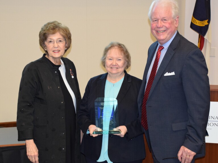 Ann Wall, a woman with blond hair, wears a teal shirt and black jacket and is holding her award. She stands with Elaine Marshall to her left and Bain Jones to the right.
