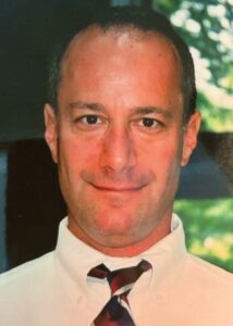 Robert, a white man with dark brown hair, is pictured outside wearing a white shirt and dark brown tie.