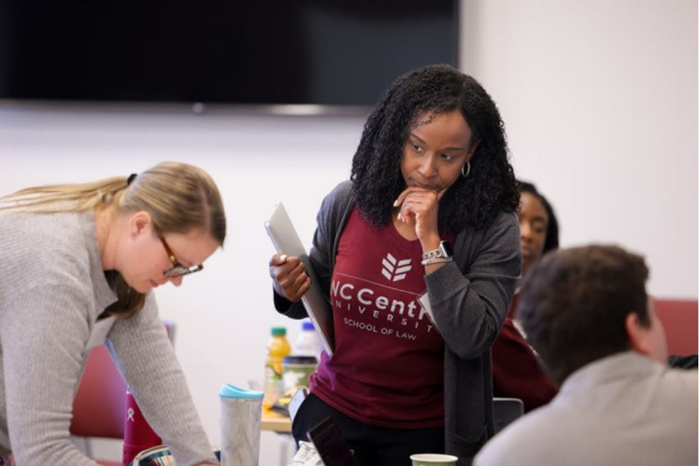 Diane, a Black woman with black hair, wears a burgundy NCCU law shirt and is pictured standing near a table where students are gathered together working. 