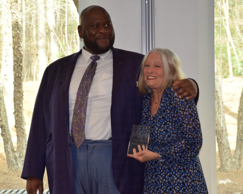 Deborah wears a blue dress with small yellow and navy dots and holds the award. Salim wears a white shirt, grey tie and dark jacket and has his arm around Deb's shoulder.