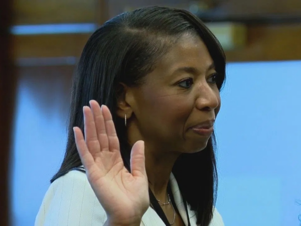 Judge Jackie Grant, a Black woman with black hair, is pictured wearing an ivory suit and is raising her right hand while being sworn in.