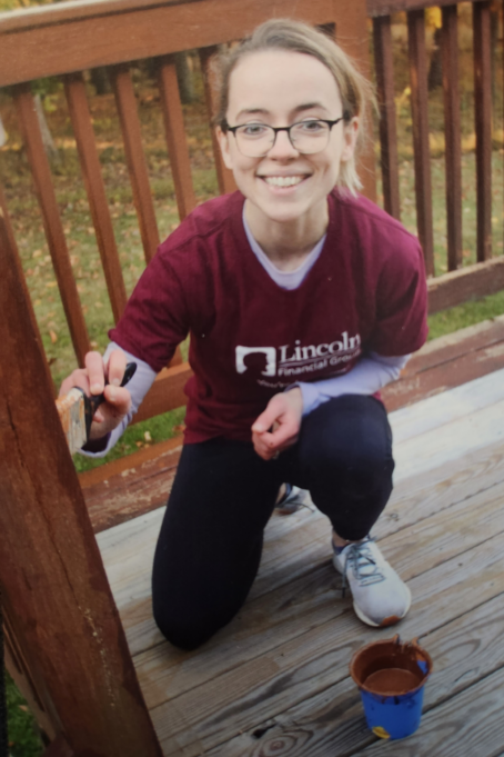Katie, a white woman with blond hair and glasses, wears a burgundy shirt and black pants is pictured painting a deck.