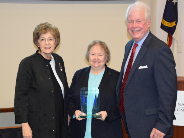 Ann Wall, a woman with blond hair, wears a teal shirt and black jacket and is holding her award. She stands with Elaine Marshall to her left and Bain Jones to the right.