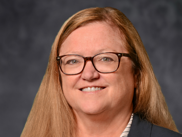 Elizabeth Croom has shoulder-length blond hair, brown glasses and wears a white blouse, navy jacket and gold circle pin.