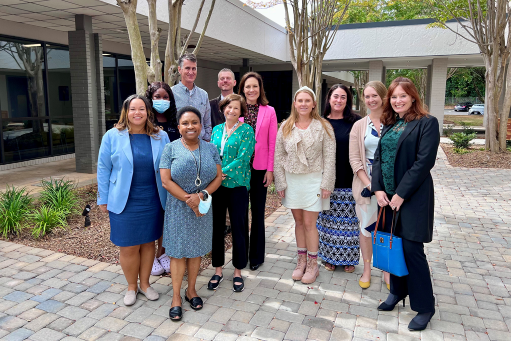 Larissa wears a blue dress and light blue jacket. Nine staff members stand with her, and Ashley stands in the middle.