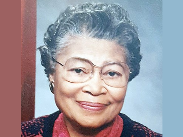 Annie Brown Kennedy, a Black woman with grey hair and clear glasses, wears a coral shirt and black sweater, and is smiling.