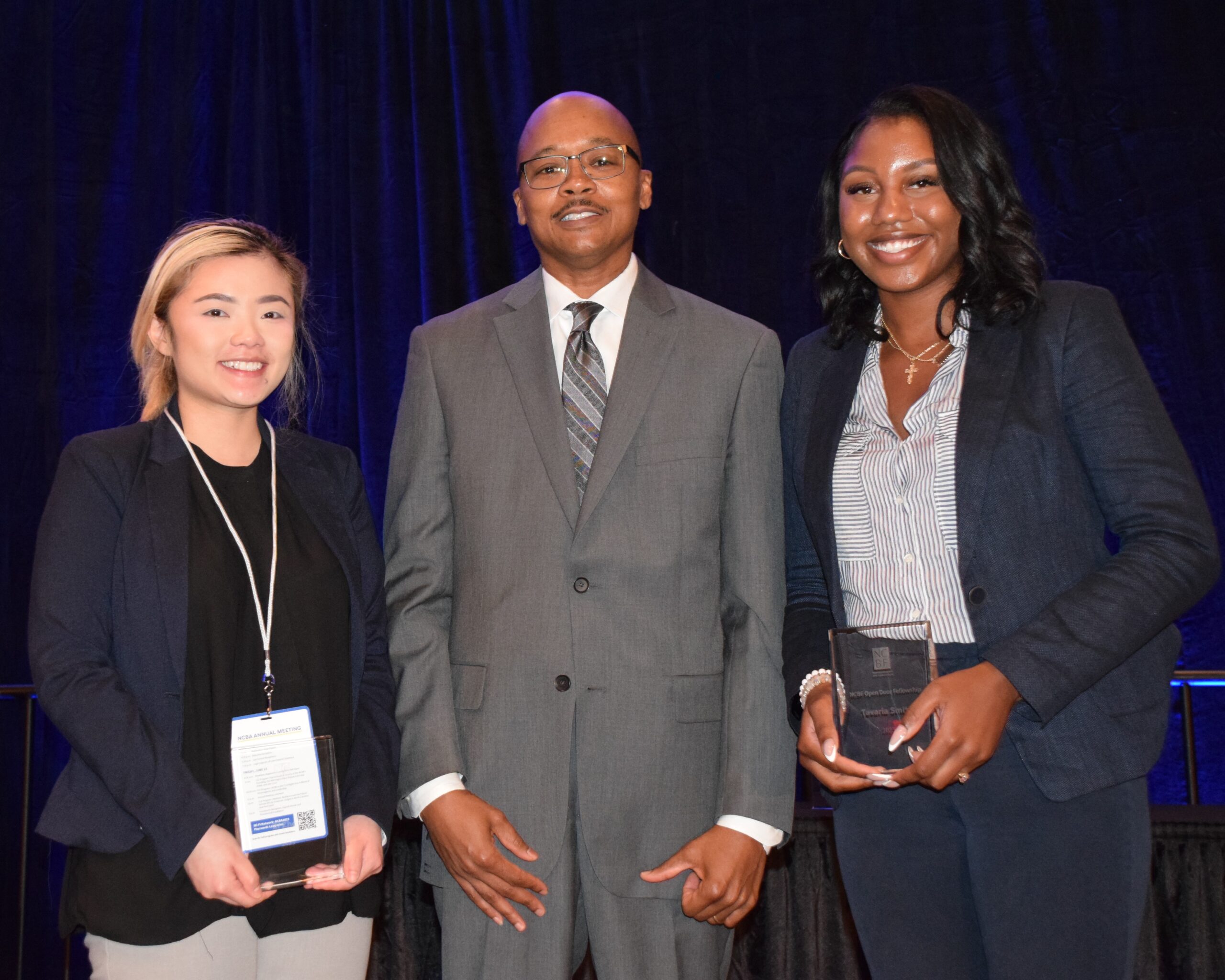 Zi, an Asian American woman with blond hair, wears a black blouse and navy jacket, Clayton, a Black man with a shaved head, wears a grey suit, and Tavaria, a Black woman with black hair, wears a white blouse and dark grey pantsuit.