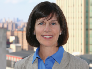Marjorie, a white woman with brown hair, wears a blue button-down shirt and an ivory jacket, and she is pictured smiling.