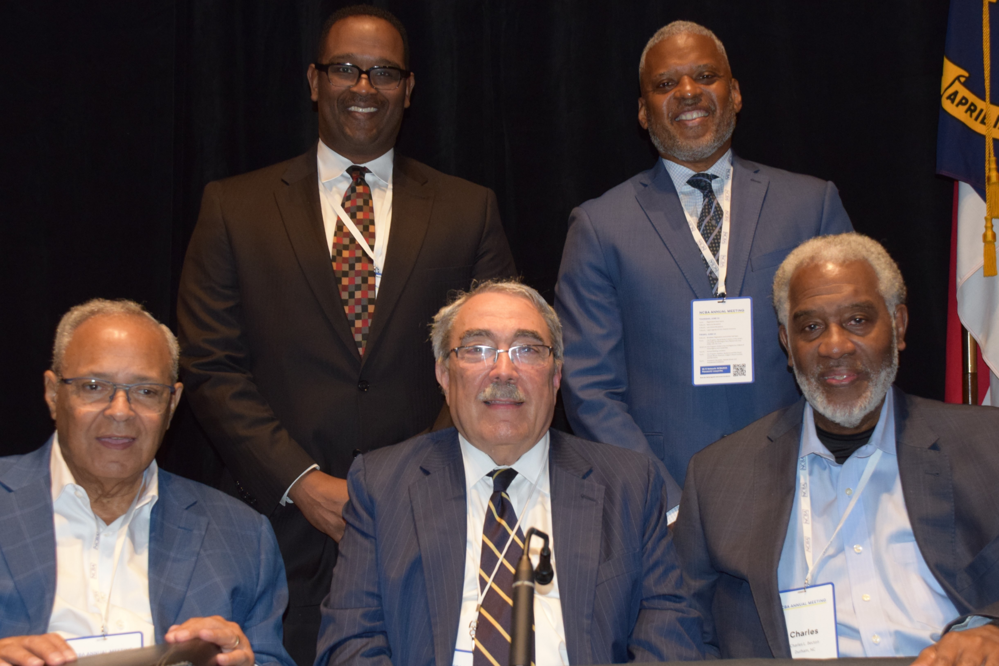 The five panelists are pictured seated, and each is wearing formal attire and smiling. Michaux is a Black man with white hair and is wearing a white shirt and grey suit. Butterfield is a Black man with grey hair and is wearing a white shirt, tie, and grey suit. Becton is a Black man with white hair and a white beard who is wearing a white shirt and black suit.