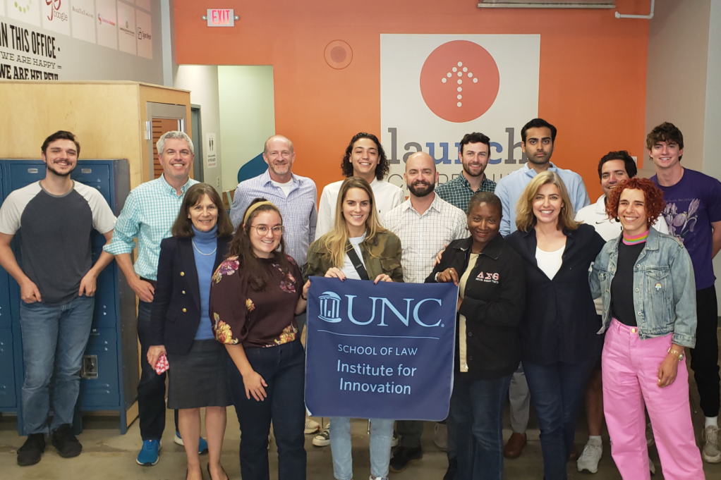 Fifteen people, including law students and faculty, are pictured in a brightly lit room. White, who stands to the left, is a white woman with brown hair and is wearing a blue shirt and black jacket and skirt. A student in the center holds a sign that reads "UNC School of Law Institute for Innovation."