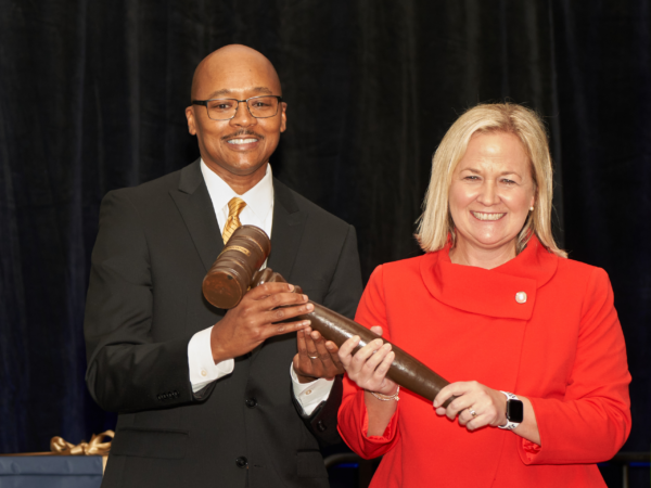 Clayton, left, a Black man with a shaved head, wears a white shirt and yellow tie. He hands the gavel to Kimberly, a white woman with blond hair who is wearing a red blouse. They are smiling.