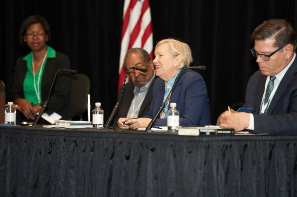 Dr. Roseboro is a Black woman with black hair wearing a green blouse and black suit. Panlists Grear, a Black man in a grey shirt and black suit, Inman, a woman with blond hair, and Paschal, a white man with brown hair, are seated.