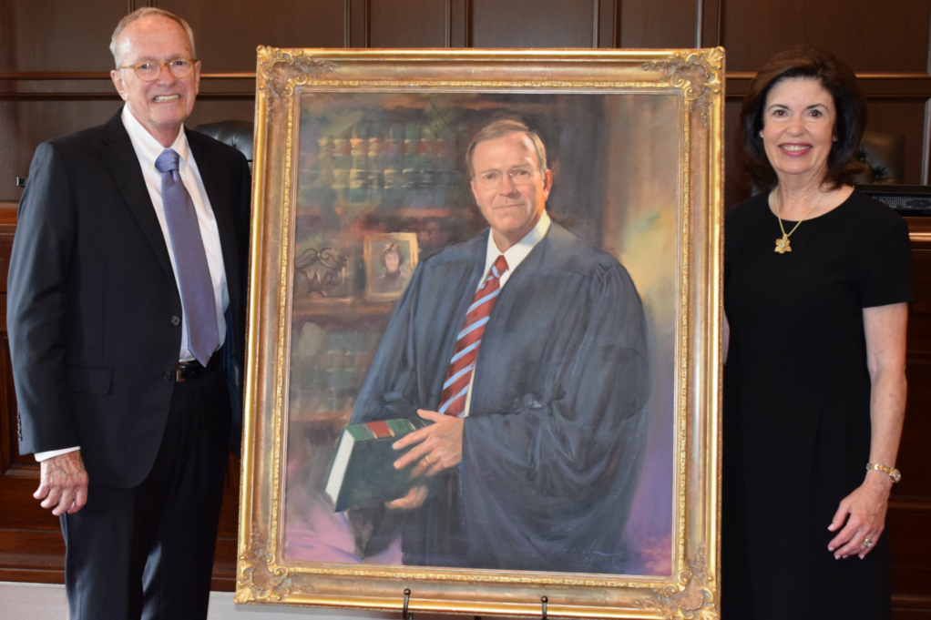 Judge Martin has brown hair and glasses and wears a white shirt, purple tie, and black suit, and Margaret, who has dark brown hair, wears a black dress. The portrait is inbetween Judge Martin and Margaret. In the portrait, Judge Martin is depicted in his judicial robe wearing a white shirt and red and blue striped tie and holding a legal book, with legal books in the background. 