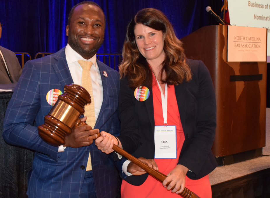 Jonathan, a Black man with black hair, wears a white shirt, yellow tie, and navy suit, and he accepts the gavel from Lisa, a white woman with brown hair wearing a coral dress.