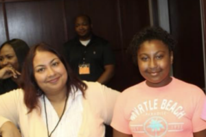Elysia is photographed standing with her daughter, Saliyah. Elysia is a woman with shoulder-length brown hair and wears a white shirt and ivory sweater. She is smiling. Her daughter, Saliyah, has dark brown hair and stands to her right. Saliyah is wearing a coral shirt that says "Myrtle Beach."