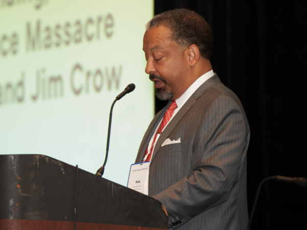 Rob Harrington, a Black man with Black hair and a beard, wears a white shirt, red tie, and grey suit jacket and stands at a podium with a PowerPoint behind him that reads "Racist Roots: A History of the NCBA’s Founding, the Wilmington Race Massacre and Coup d’état, and Jim Crow."