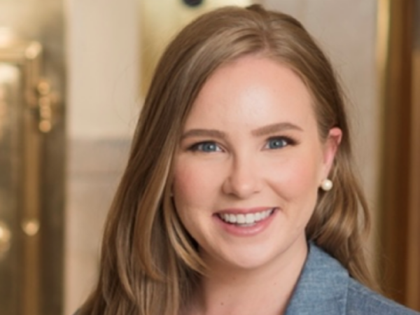 Elizabeth Brooks Savage, a white person with long auburn hair, wears an ivory blouse with a chambray-colored blazer and pearl earrings.