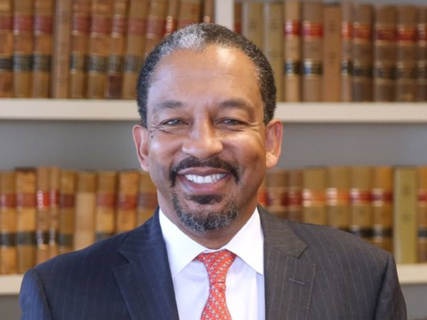 Rob Harrington, a Black man with black hair and a beard, wears a white shirt, red tie, and black suit, and stands in front of a bookshelf with red and gold books.