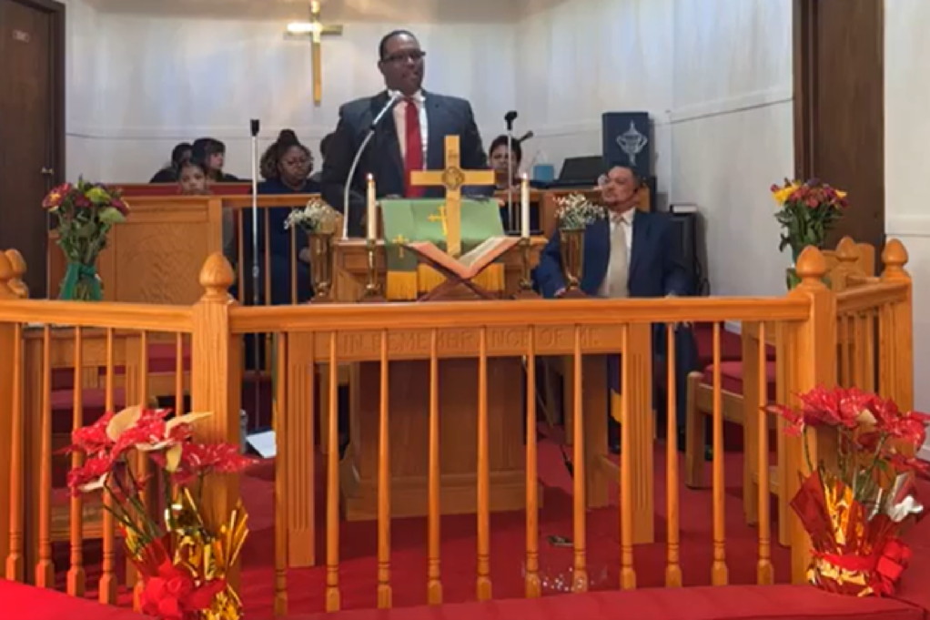 Ted, a Black man with black hair, wears a white shirt, red tie, and black glasses. He is standing behind a wooden pulpit. The pulpit has a cross on the center. A choir sits in pews behind him.