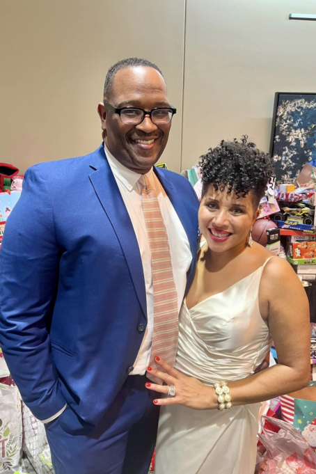Ted, a Black man with black hair, wears a white shirt, navy suit, and peach and off-white tie. He is pictured with his spouse, Tiffany, who is a Black woman with curly black hair. She is wearing an off-white satin dress.