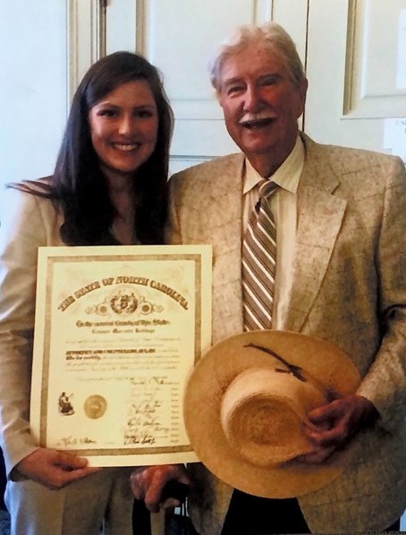 Horton, a white man with white hair and a mustache, wears a light brown suit, off-white shirt and brown and off-white striped tie. He holds an off-white hat. His granddaughter Eleanor stands to the left, and she is a white woman with brown hair. She is wearing an off-white pantsuit.