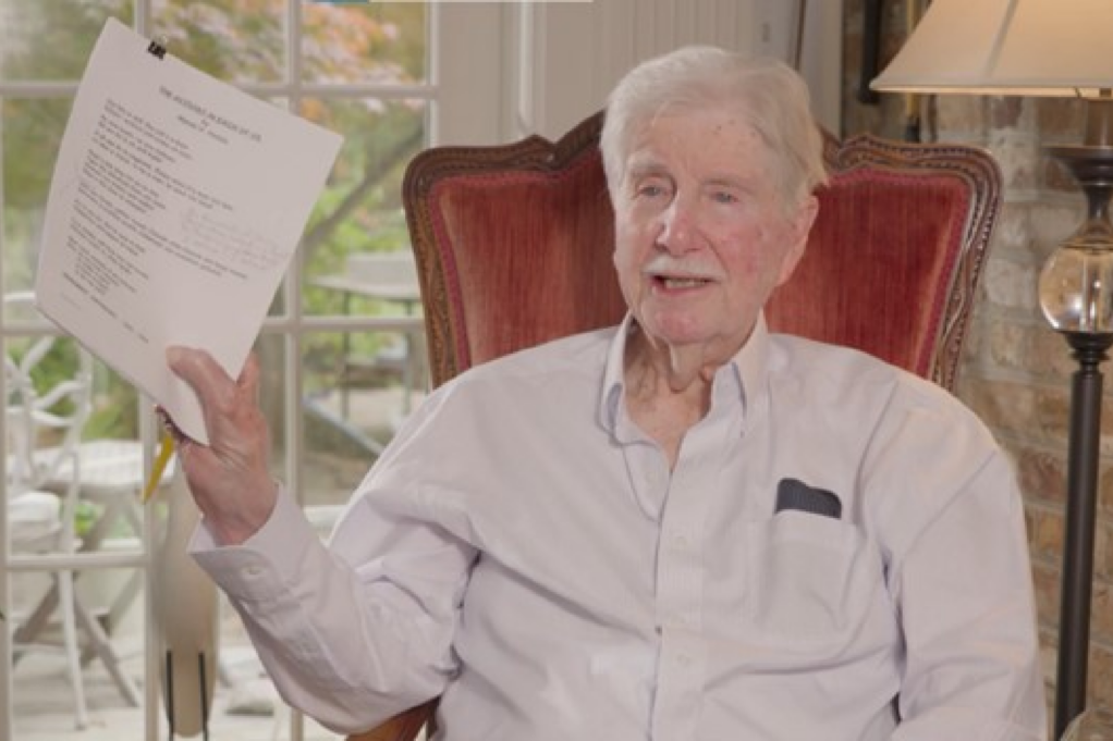 Horton, a white man with white hair, wears a pale grey shirt and sits in a red velvet chair. He holds a paper with a poem commemorating the Kingsboro victory.