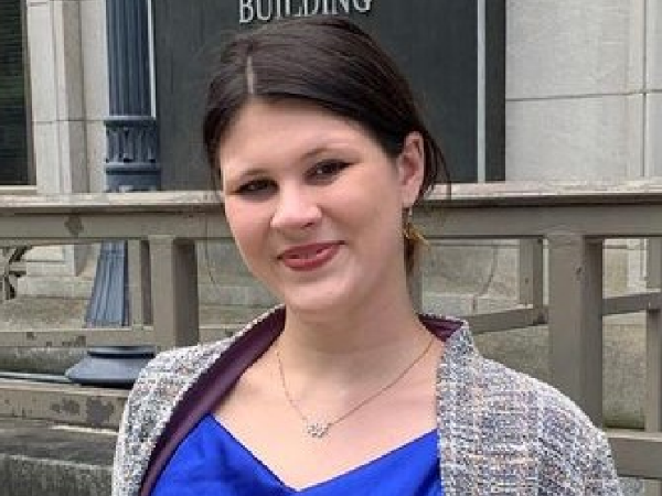 Emily, a woman with dark brown hair in a ponytail, wears a royal blue top, ivory and brown jacket, and silver necklace. She is pictured outside a courthouse.