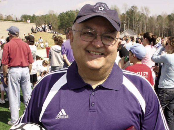 Woody, a white man with grey hair, wears a purple and white Adidas polo and a purple hat with the image of a soccer ball that says "soccer." A soccer field and onlookers are behind him.