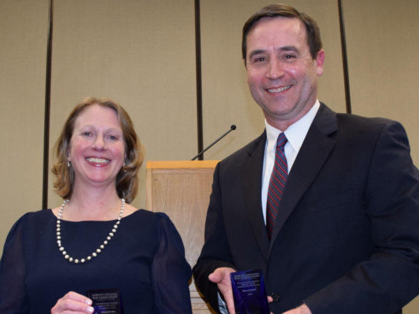 Martha, a white woman with light brown hair, wears a navy dress and pearl necklace, and Kearns, a white man with brown hair, wears a white shirt, red and blue striped tie, and black suit. They are holding dark blue awards.