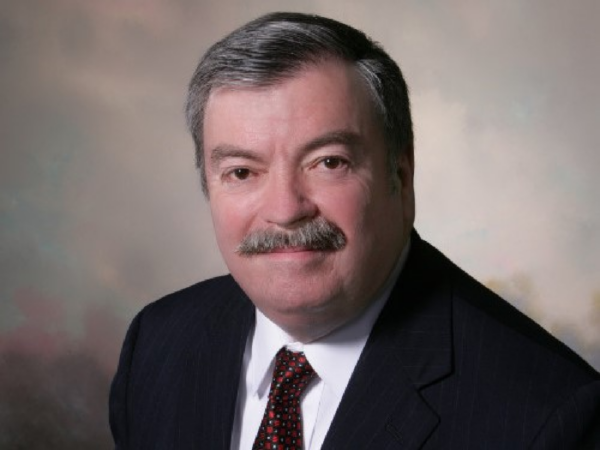 Mark, a white man with grey hair and a grey mustache, wears a white shirt, red tie, and black suit.