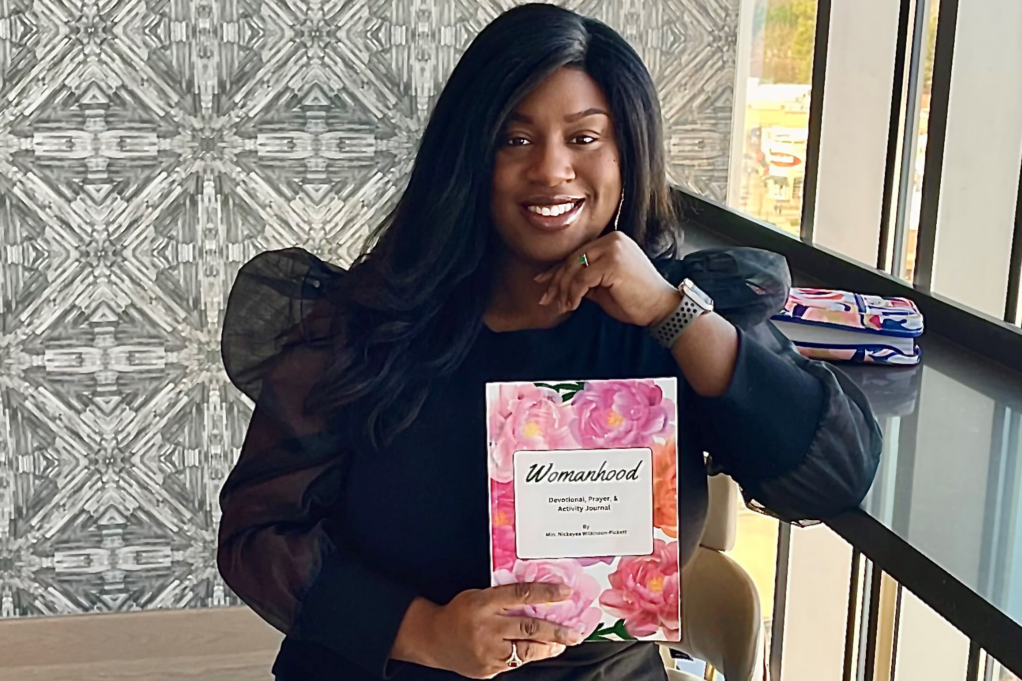 Nicky, a Black woman with long clack hair, wears a black blouse with ruffled sleeves. She holds a book called "Womanhood: A Devotional Prayer & Activity Journal." The book cover is her own art and consists of bright pink flowers.