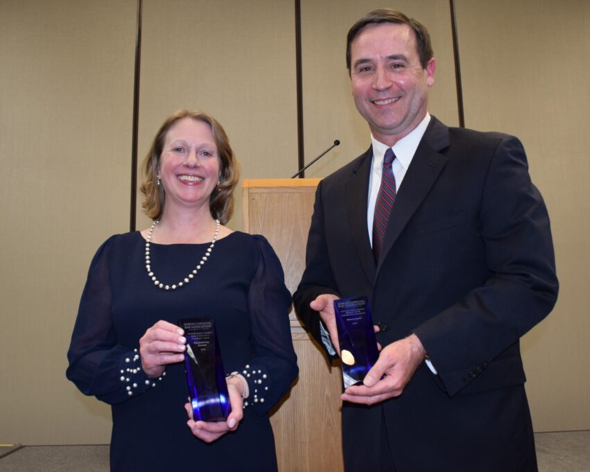 Martha, a white woman with light brown hair, wears a navy dress and pearl necklace, and Kearns, a white man with brown hair, wears a white shirt, red and blue striped tie, and black suit. They are holding dark blue awards.