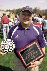 Woody, a white man with grey hair, wears a purple and white Adidas polo and a purple hat with the image of a soccer ball that says "soccer." A soccer field and onlookers are behind him.