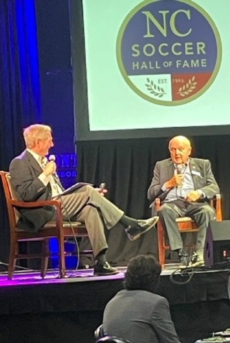 Woody sits on a chair onstage during the NC Soccer Hall of Fame. He wears a pale blue shirt and grey suit. 