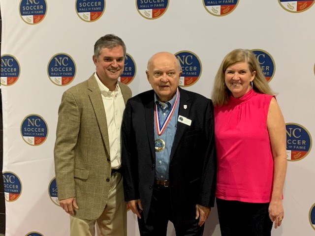Woody, a white man with a shaved head, wears a blue shirt and black suit and a medal from the NC Hall of Fame. The medal is gold and the ribbon around it is red, white, and blue. He stands with son Reggie, a white man in a brown suit, and daughter Elizabeth, a white woman in a coral tank top.