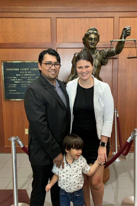 Almanza, a woman with dark brown hair, wears a black dress and white blazer. Her spouse, a man with dark brown hair and black glasses, wears a suit and grey shirt. They hold hands with their little boy, about three years old, who has dark hair and wears a white shirt with dinosaurs.