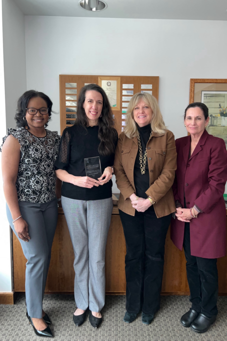 Jen, a white woman with long brown hair, wears a black blouse, grey pants and black shoes. She stands with LaToya, a Black woman with black hair and glasses, who wears a black and white flowered shirt and grey pants, Michelle, a white woman with blond hair who wears a black shirt, black pants, and tan jacket, and Stacey, a white woman with brown hair who wears a black blouse and pants and a maroon jacket.