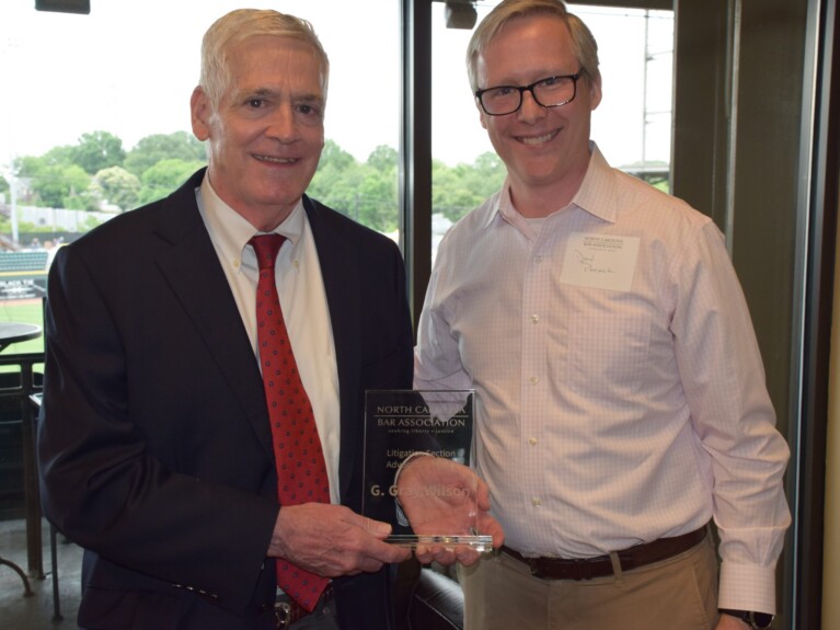 Gray Wilson, a white man with white hair, wears a white shirt, red tie with black dots, and black suit. Don Pocock, a white man with blond hair and brown glasses, wears a white shirt with khaki pants.