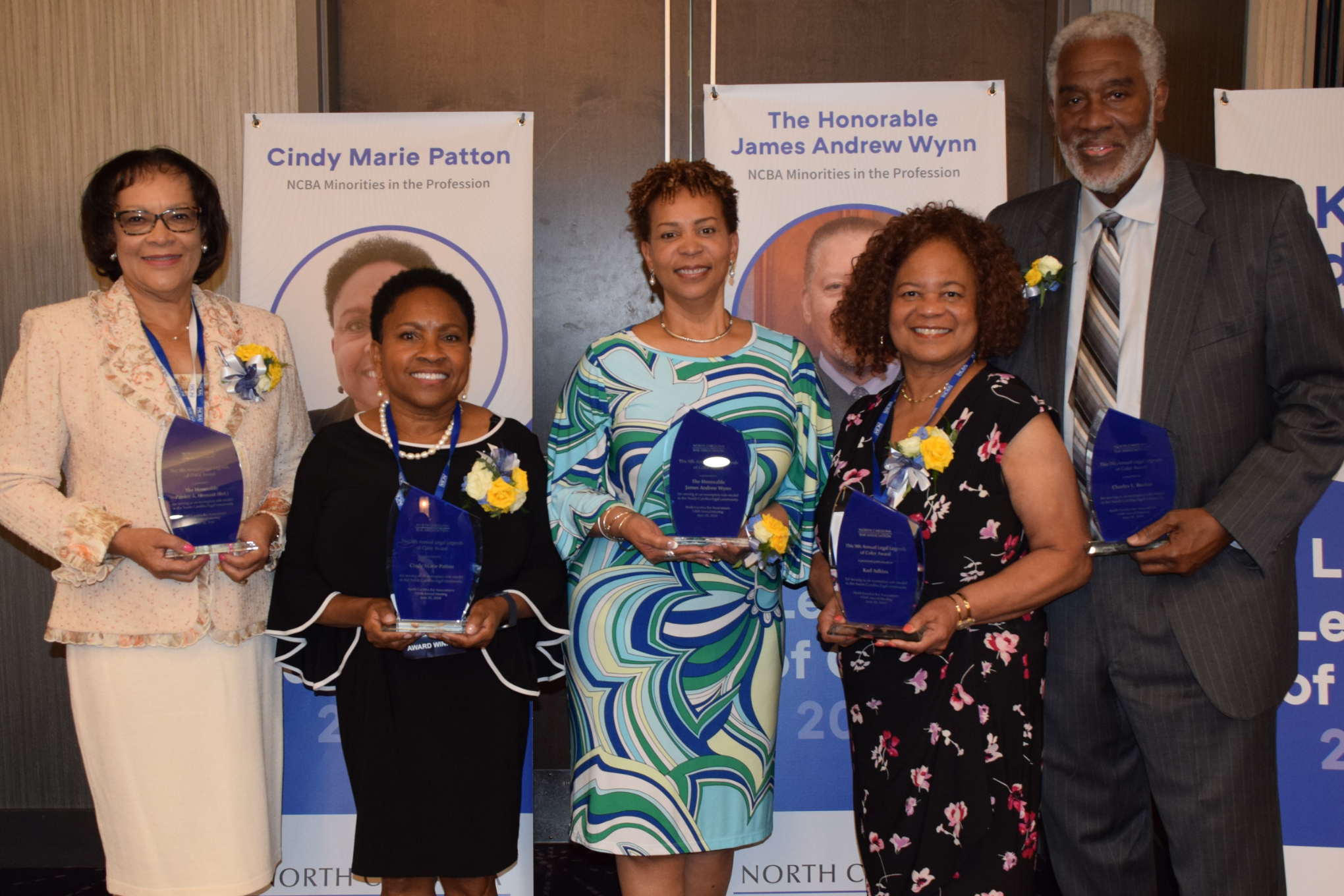 The 2024 Legal Legends of Color and their representatives are pictured holding their awards. Judge Hinnant is a black woman with black hair and is wearing a peach dress and blazer, Cindy Patton is a Black woman with black hair wearing a black dress, Judge Karlene Turrentine is a Black woman with brown hair wearing a green and blue dress, Carriette Adkins is a Black woman with brown hair wearing a black dress with pink flowers, and Charles L. Becton is a Black man wearing a grey shirt, grey and brown striped tie, and grey pinstripe suit.