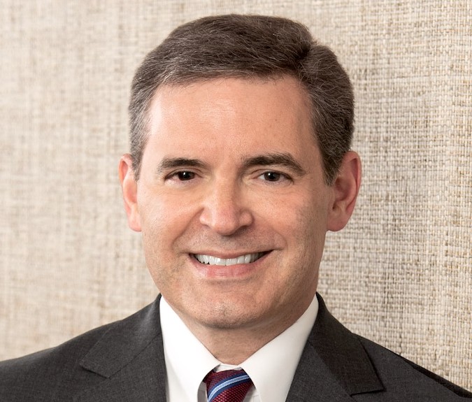 John Cella is a man with brown hair. He wears a white shirt, dark grey suit and maroon and blue striped tie.