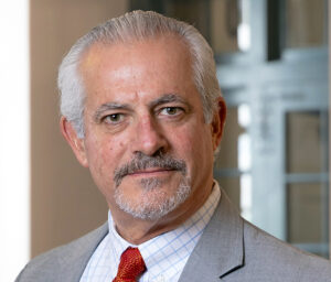 Peter Juran is a man with grey hair and a beard and mustache. He wears a white shirt, red tie and light grey suit.