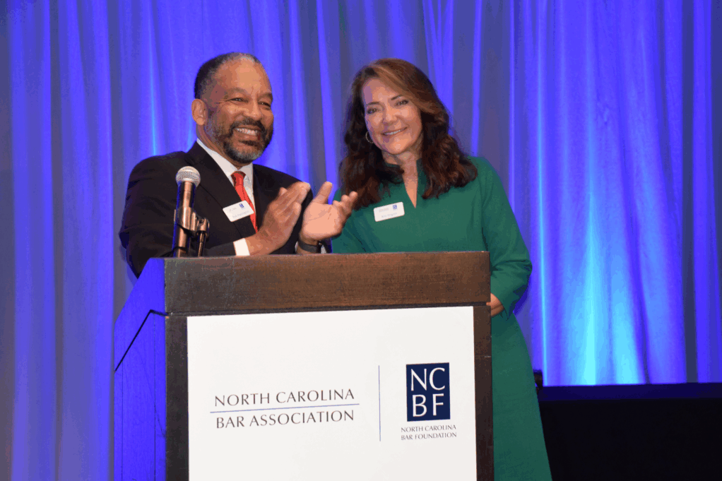 Rob, a Black man with black hair, wears a white shirt, red tie and black suit. Kim, a white woman with brown hair, wears a green dress.