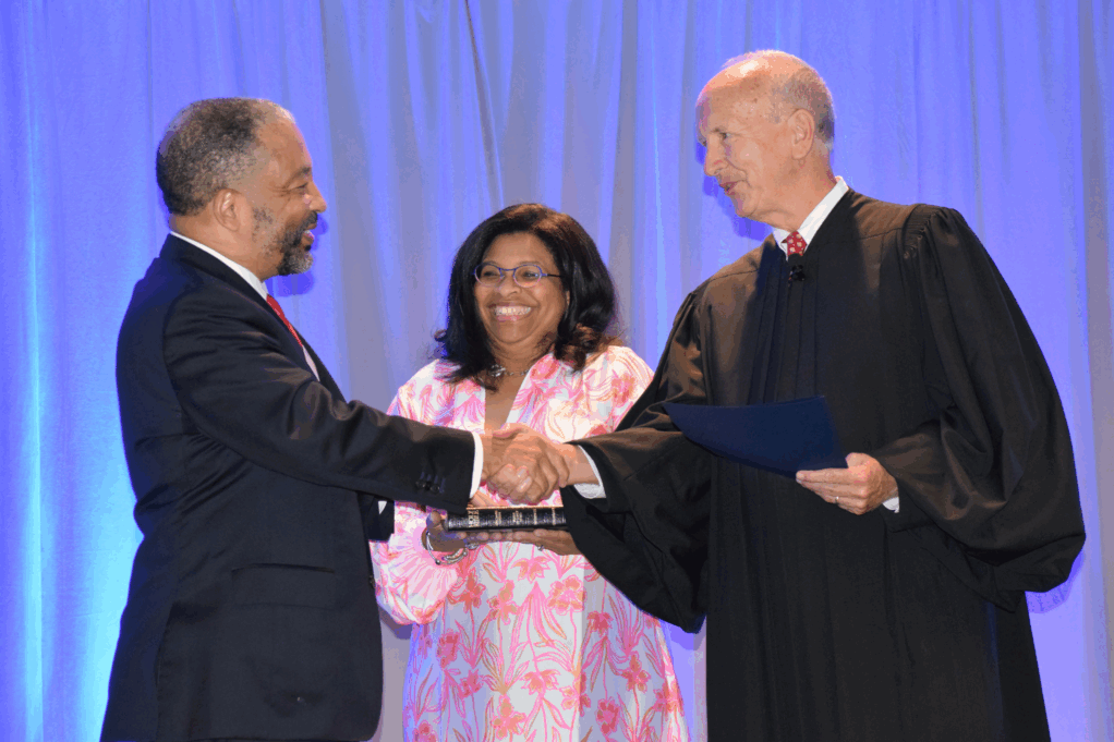 Sharon is a Black woman with black hair and is wearing a pink dress. Rob wears a black suit and is shaking hands with Newby, a white man with grey hair.