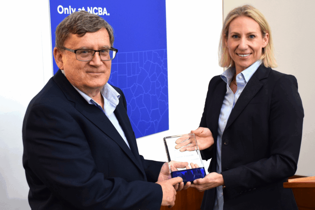 Archer, a white man with brown hair, wears a light blue shirt and black suit. Smaxwell, a white woman with blond hair, wears a light blue shirt and black suit.