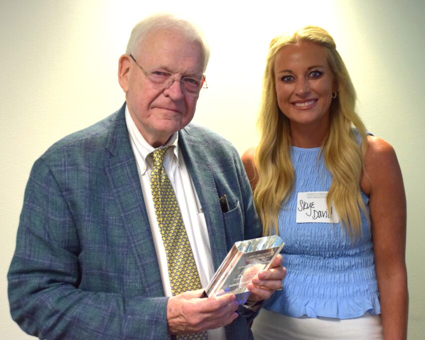 Borden Parker, a white man with white hair, wears a white shirt, yellow tie and blue suit. David, a white woman with blond hair, wears a light blue blouse.