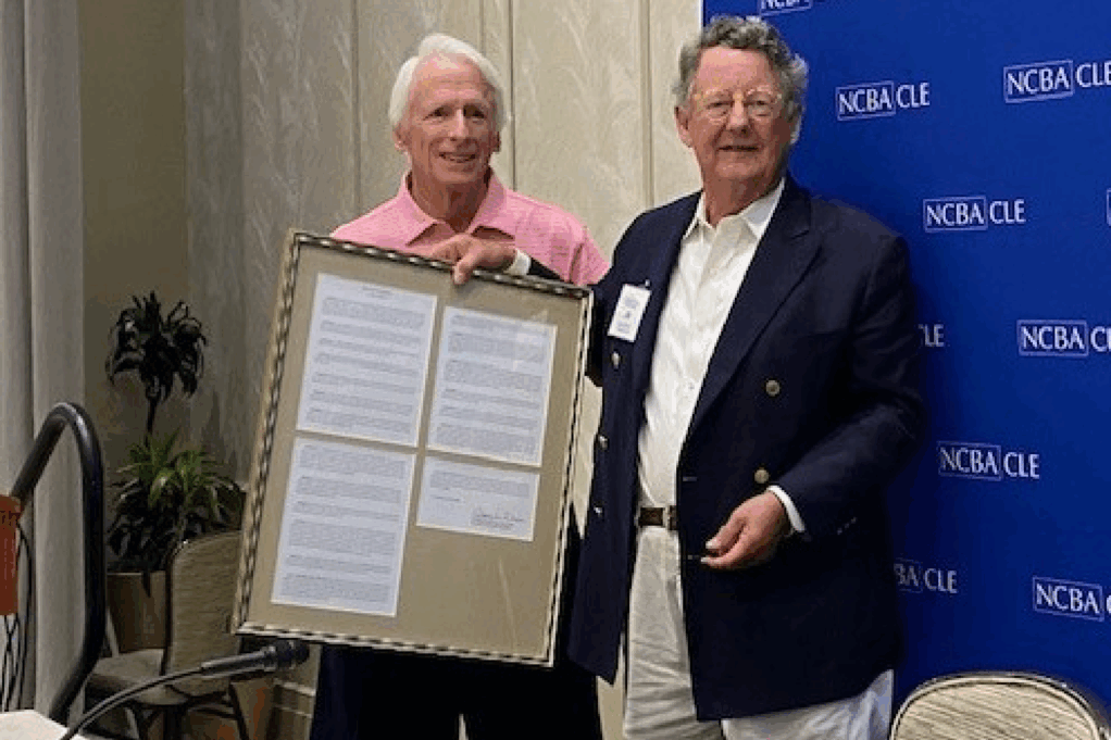 Johnsey, a white man with white hair, wears a coral shirt, and Narron, a white man with grey hair, wears a white shirt and navy blue jacket. 