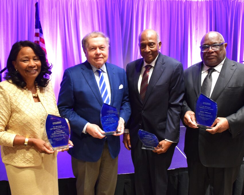 The four dignitaries are pictured holding their awards. Copeland Biggs is in a yellow dress and jacket, John Wester wears a blue jacket, white shirt and blue and white striped tie, Edison wears a black suit and dark red tie, and Williams wears a black suit and black tie.