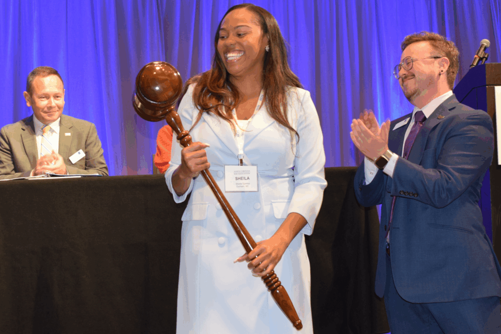 Sheila, a Black woman with brown hair, wears a light grey dress and jacket. She holds a large brown gavel. Collins is a person with brown hair and glasses and is wearing a blue suit.