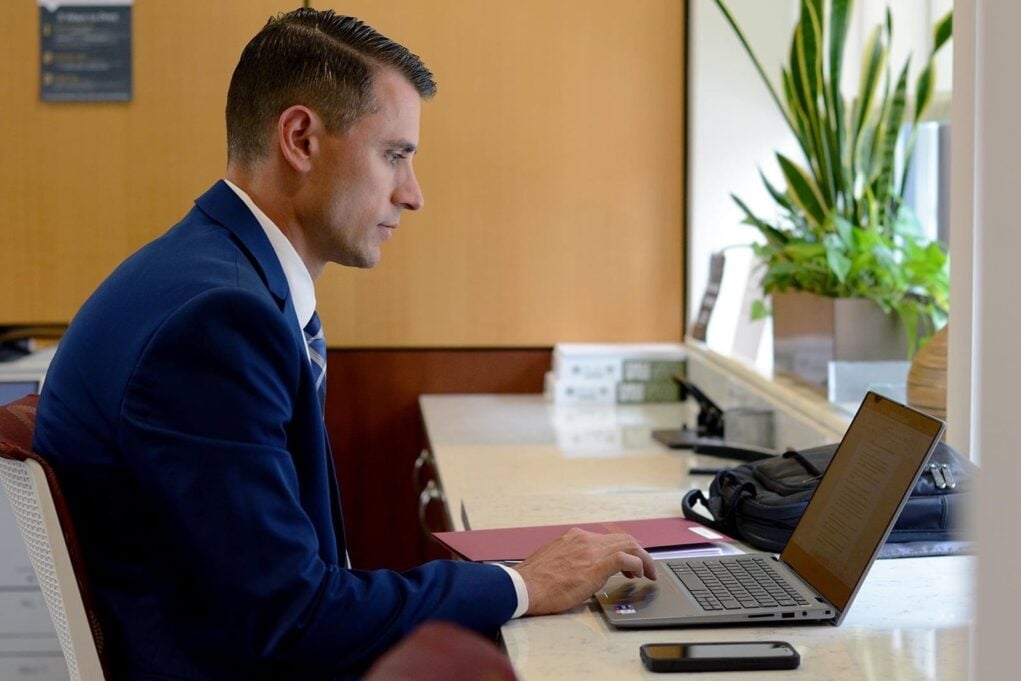Bowyer, a white man with brown hair, wears a navy blue suit, white shirt and plaid tie. He is pictured seated at a coffee bar working on his laptop.