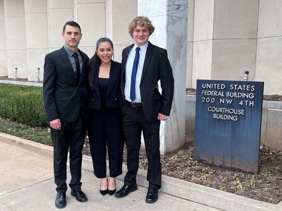 Bowyer, a white man with brown hair, wears a black suit. He is pictured with a white woman with dark hair wearing a black suit, and a white man with blond hair wearing a white shirt, blue tie and black suit. They stand outside a U.S. Federal Courthouse.